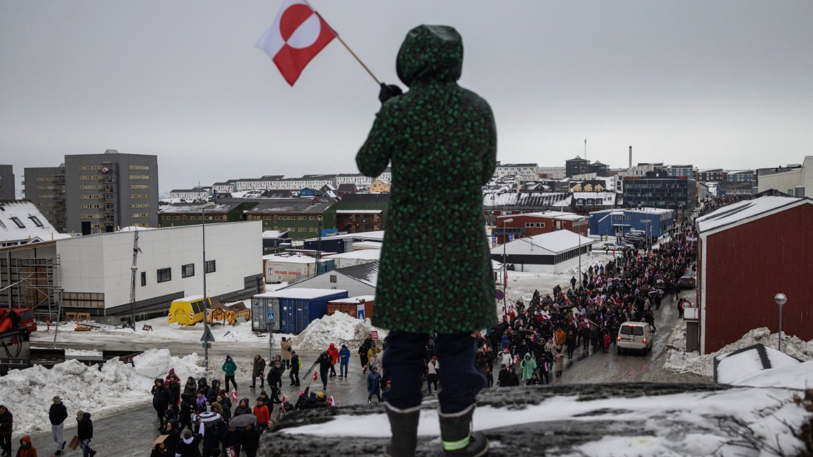 امرأة تلوّح بعلم غرينلاند  تقف على ربوة تكسوها الثلوج، بينما يشارك سكان في احتجاج، بنوك عاصمة  الجزيرة ضد مطالبة الرئيس دونالد ترامب بالتنازل عن الجزيرة القطبية الشمالية للولايات المتحدة. - Reuters