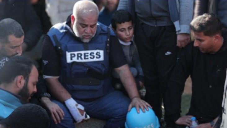 الصحفي الدحدوح مرتدياً سترة الصحفيين، يده مصابة ويجلس حزيناً بين جمع من الناس. - BBC