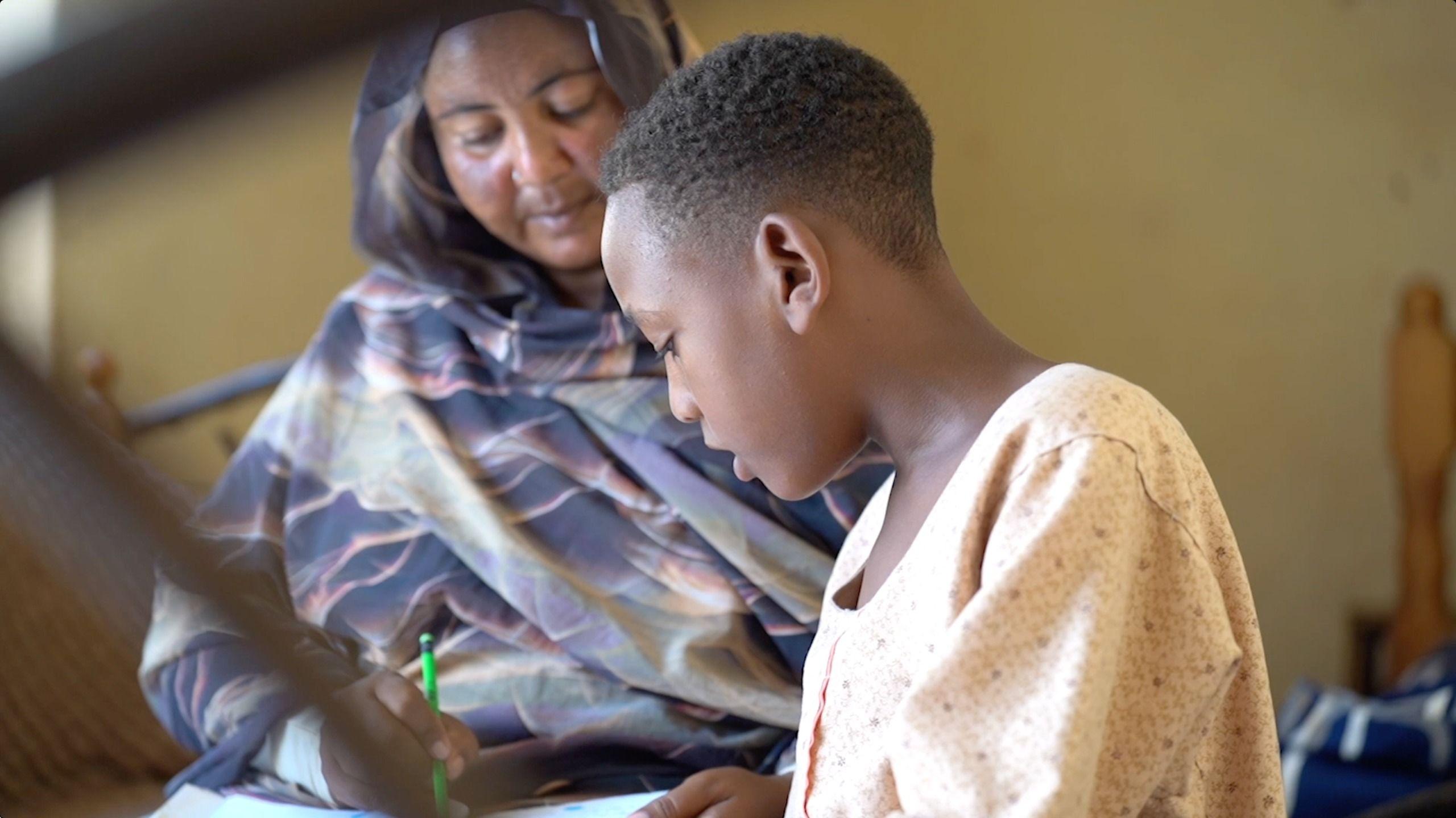 يجلس الطفل مصطفى عثمان، 10 سنوات، حزيناً، إلى جانب والدته شيراز عماد الدين، وهو تراجع له بعض الدروس. - BBC