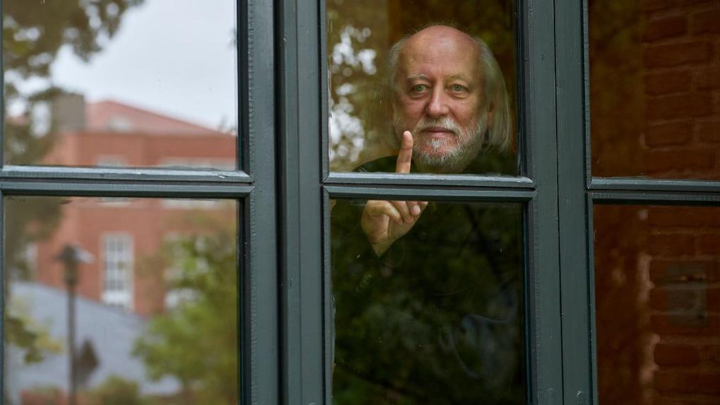 الكاتب الهنغاري لازلو كراسنهوركاي يقف خلف نافذة زجاجية، يرفع إصبعه بهدوء وينظر بتأمل إلى الخارج، فيما تنعكس خلفه ملامح مبانٍ من الطوب وأشجار خريفية. - Getty Images