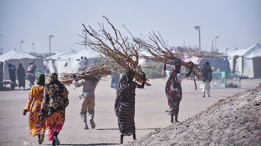 تظهر نساء سودانيات من الخلف باللباس التقليدي يحملن حطباً، وعدد آخر من الرجال يحمل أحدهم حطباً. كل شخص في الصورة يظهر ماشياً لوحده قرب عدد من الخيام. الصورة التقطت نهاراً. - Anadolu via Getty Images