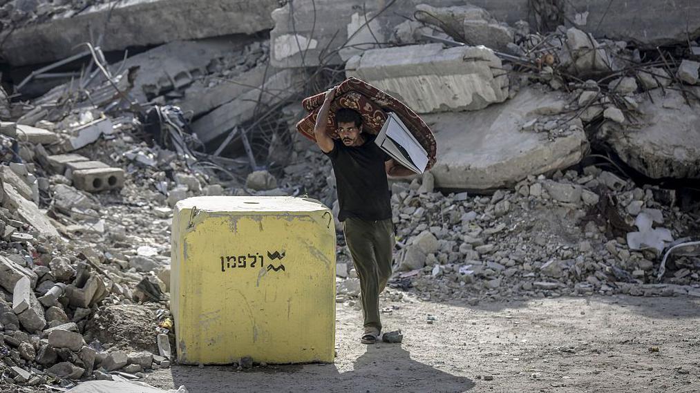 رجل يحمل مرتبة مطوية في غزة يمر بجوار كتلة خرسانية صفراء وضعها الجيش الإسرائيلي، وتظهر الأنقاض في الخلفية - Saeed M. M. T. Jaras/Anadolu via Getty Images