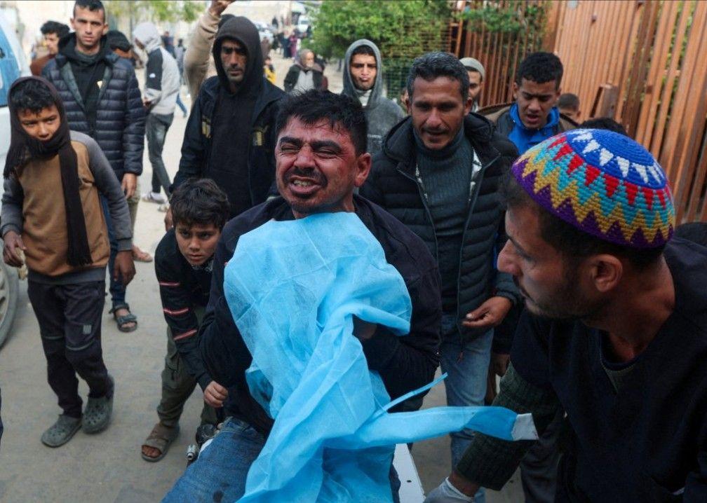الأب بشعره الأسود وبشرته السمراء، يحمل جثمان ابنه الملفوف بقماش أرزق، وحوله عدد من الرجال والأطفال الفلسطينيين. الصورة التقطت نهاراً. - Reuters