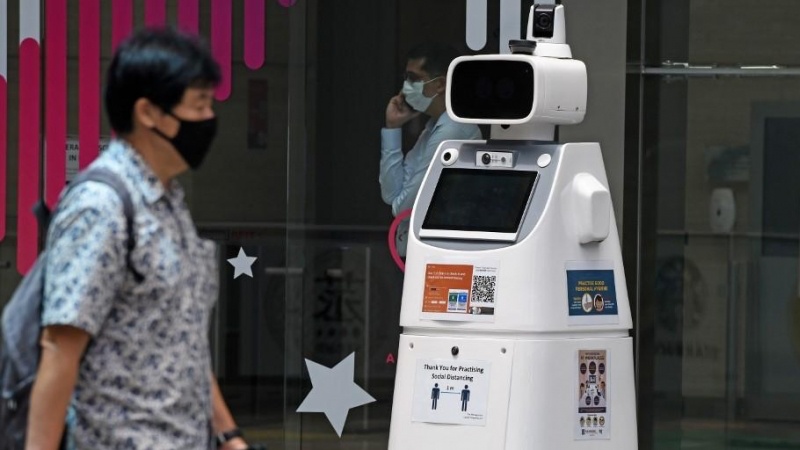 رجل يرتدي قناعًا للوجه يمر أمام روبوت مبرمج للحفاظ على التباعد الاجتماعي أمام مبنى للمكاتب في سنغافورة