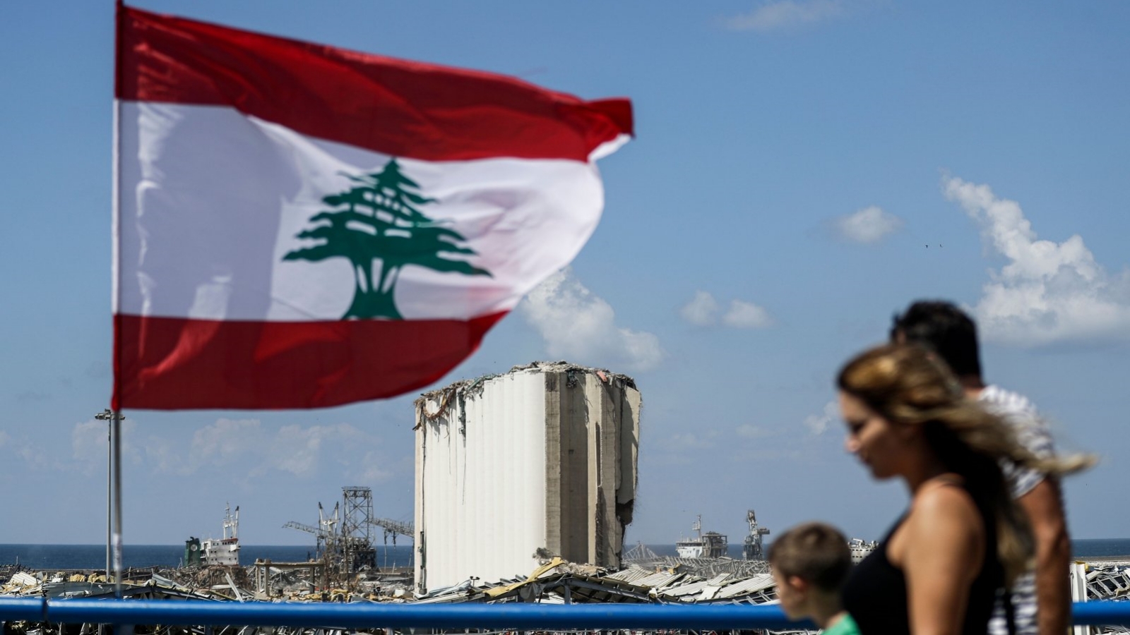 مواطنون يمشون على جسر أمام العلم اللبناني بالقرب من ميناء العاصمة اللبنانية بيروت. حيث تظهر إهراءات الحبوب المدمرة بفعل الإنفجار في الخلفية. 9 آب/أغسطس 2020.