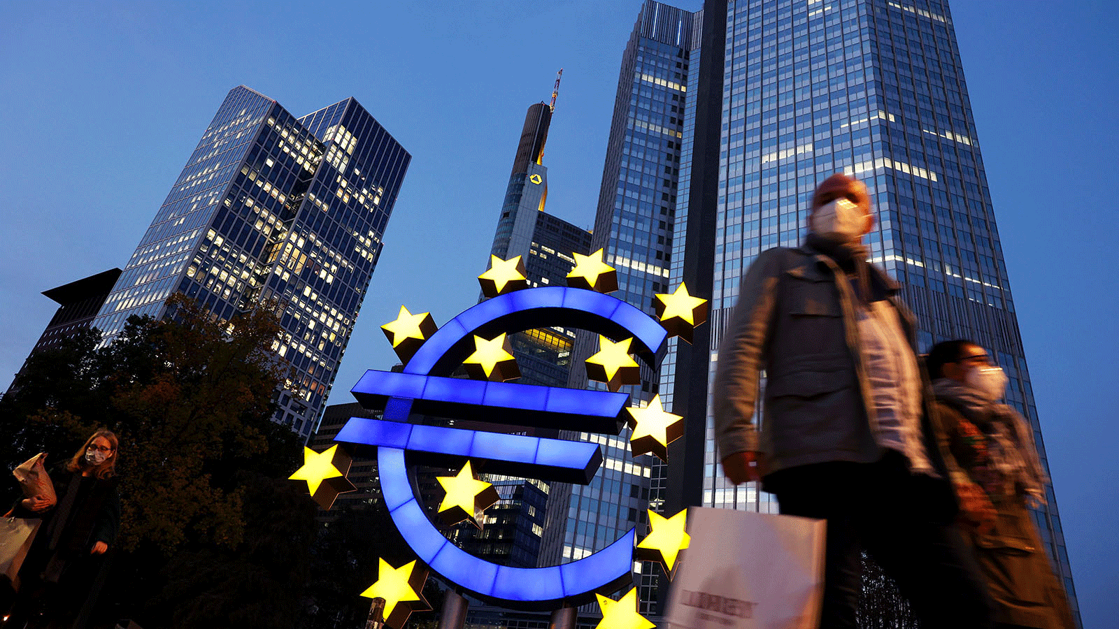أشخاص يرتدون أقنعة الوجه يسيرون أمام لافتة اليورو والمقر السابق للبنك المركزي الأوروبي في فرانكفورت، ألمانيا.