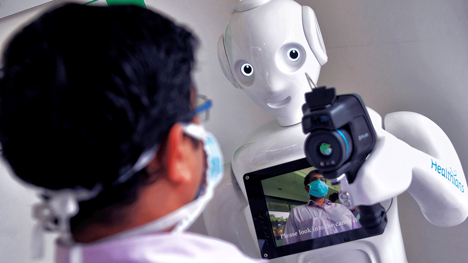  عامل في المستشفى يقف أمام روبوت مزود بكاميرا حرارية مثبتة لإجراء فحوصات أولية للمساعدة في منع انتشار COVID-19. حاليًا ،تنطبق معايير قانونية مختلفة على البشر والذكاء الاصطناعي الذين يؤدون نفس المهام.
