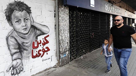 رجل وابنه يمشيان أمام جدارية مرسومة على جدار مبنى في ساحة النور في طرابلس شمالي لبنان تندد بالطبقة السياسية اللبنانية التي جوعت شعبها