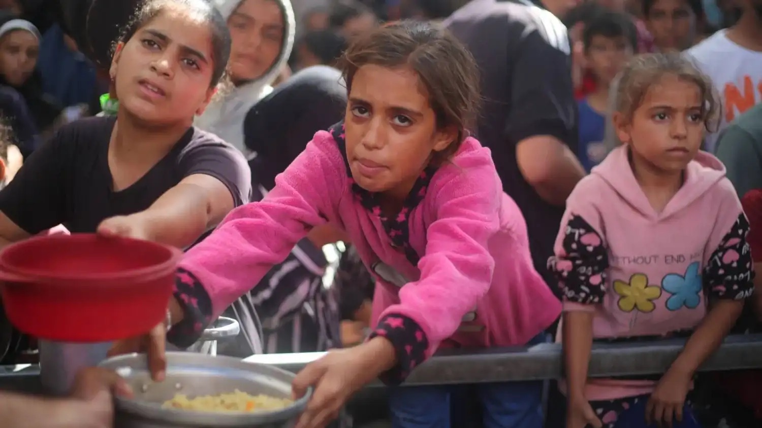 فتاة فلسطينية تمد يديها بوعاء لتلقّي حصة طعام من إحدى التكايا الخيرية بمخيم النصيرات وسط قطاع غزة