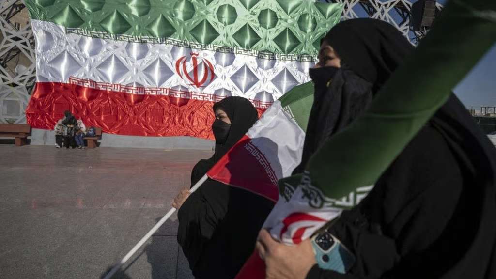 صورة من طهران – 30 كانون الأول (ديسمبر) 2025: امرأتان إيرانيتان ترتديان الحجاب تحملان الأعلام الوطنية أثناء مرورهما قرب علم إيراني ضخم، خلال مشاركتهما في مسيرة مؤيدة للحكومة في جنوب طهران