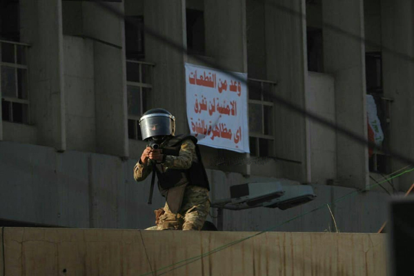 أحد عناصر القوات الحكومية العراقية يواجه المتظاهرين ببندقية صيد وخلفه لافتة لها تعد بعدم الاعتداء عليهم