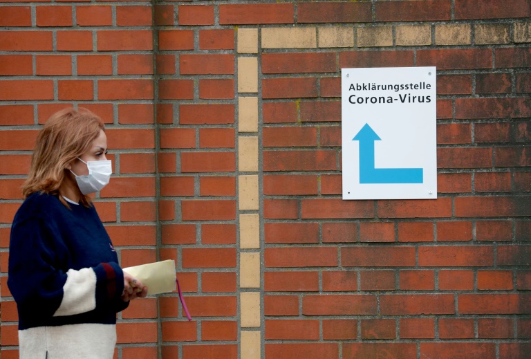 امرأة ترتدي قناعاً تعبر أمام لافتة لمركز إجراء فحوصات فيروس كورونا المستجد في برلين في 13 آذار/مارس 2020