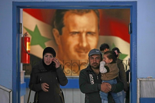 رجل يحمل طفله يقف مع سيدة عند مدخل أحد أقسام مستشفى في دمشق في 19 مارس 2020 وتبدو خلفهم صورة للرئيس بشار الأسد على العلم السوري