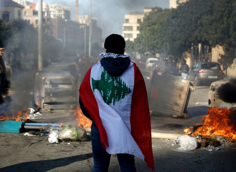 متظاهر يلف نفسه بالعلم اللبناني ويقف وسط طريق مقطوع بالأطر المشتعلة ومستوعبات النفايات، في بيروت في 14 كانون الثاني/يناير 2020