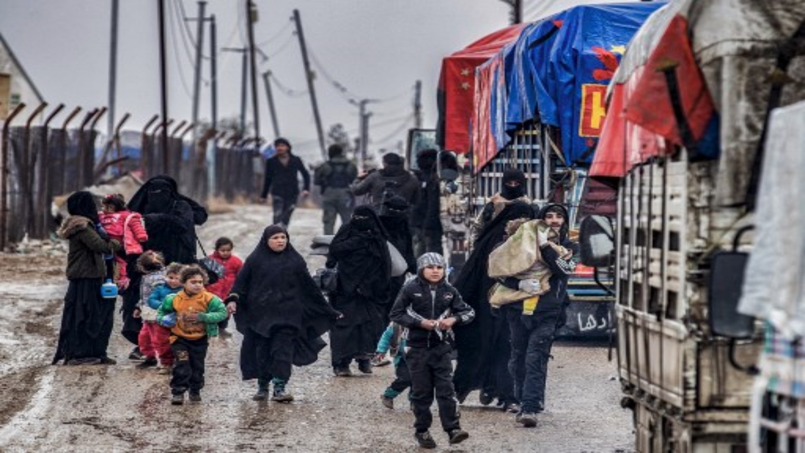 رجل يحمل طفلًا وهو يسير مع نساء وأطفال آخرين أمام أحد عناصر جهاز الأمن الداخلي الكردستاني السوري المعروف باسم الأسايش أثناء توجههم نحو الشاحنات ، أثناء إطلاق سراح الأشخاص المشتبه في ارتباطهم بمقاتلي تنظيم الدولة الإسلامية (داعش) من مخيم الهول الذي يديره الأكراد في محافظة الحسكة شمال شرق سوريا ، في 19 (يناير) 2021
