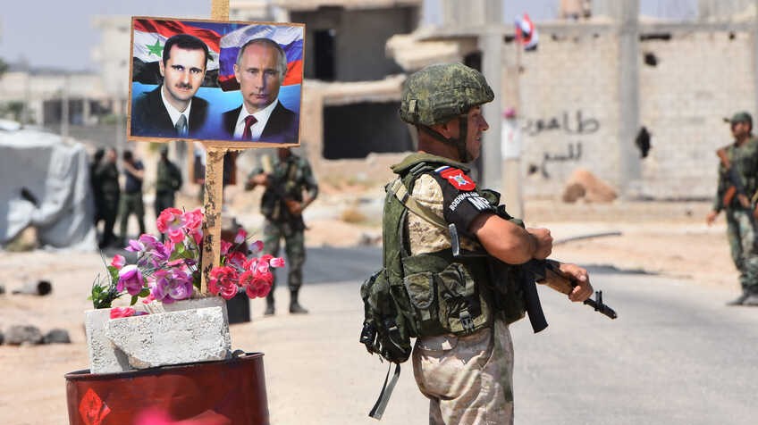 عنصران روسي وسوري في سوريا أمام ملصق يضم صورتي الرئيسين الروسي فلاديمير بوتين والسوري بشار الأسد