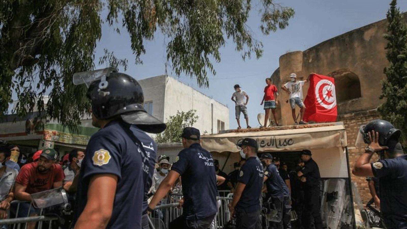 أنصار قيس سعيّد يرفعون العلم التونسي على سطح متجر أمام شرطة مكافحة الشغب، خلال مظاهرة أقيمت أمام مبنى البرلمان التونسي في باردو ، بالعاصمة تونس. بتاريخ 26 تموز/ يوليو 2021.