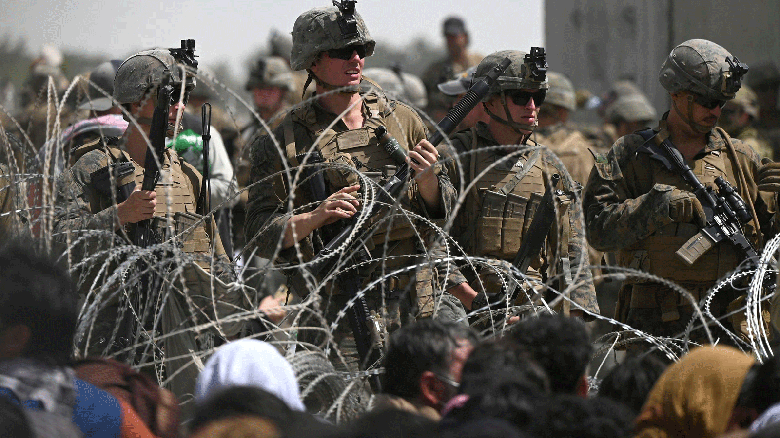جنود أمريكيون يقفون خلف الأسلاك الشائكة بينما يجلس الأفغان على جانب طريق بالقرب من الجزء العسكري من مطار كابول في 20 آب/ أغسطس 2021، على أمل الفرار من البلاد بعد سيطرة طالبان العسكرية على أفغانستان