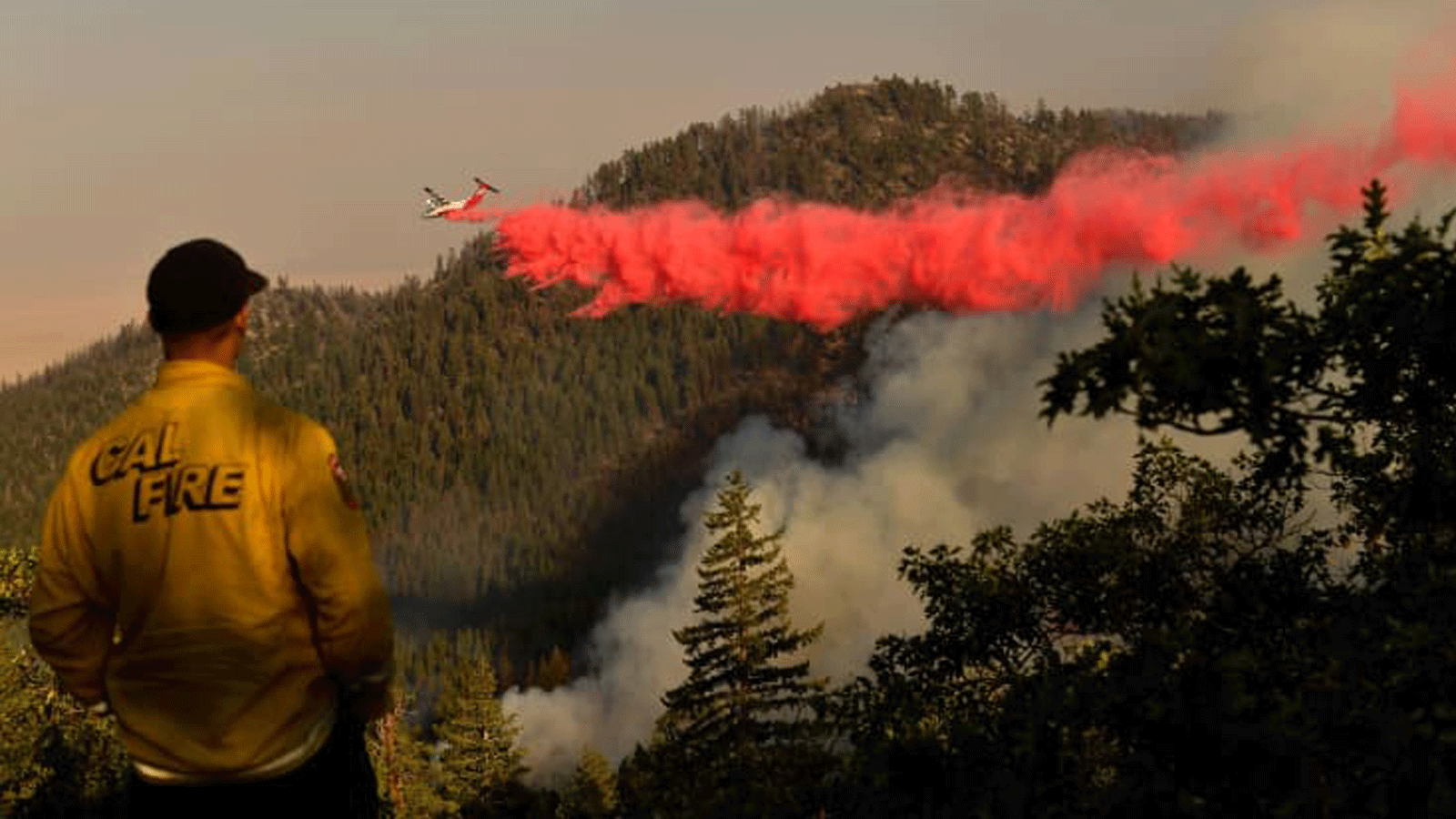 رجل إطفاء من Cal Fire من وحدة Lassen-Modoc يراقب بينما تقوم ناقلة جوية بإسقاط مثبطات الحريق على نار ديكسي حيث تحترق الأشجار على أحد التلال في 18 آب/ أغسطس 2021 بالقرب من جانيسفيل، كاليفورنيا