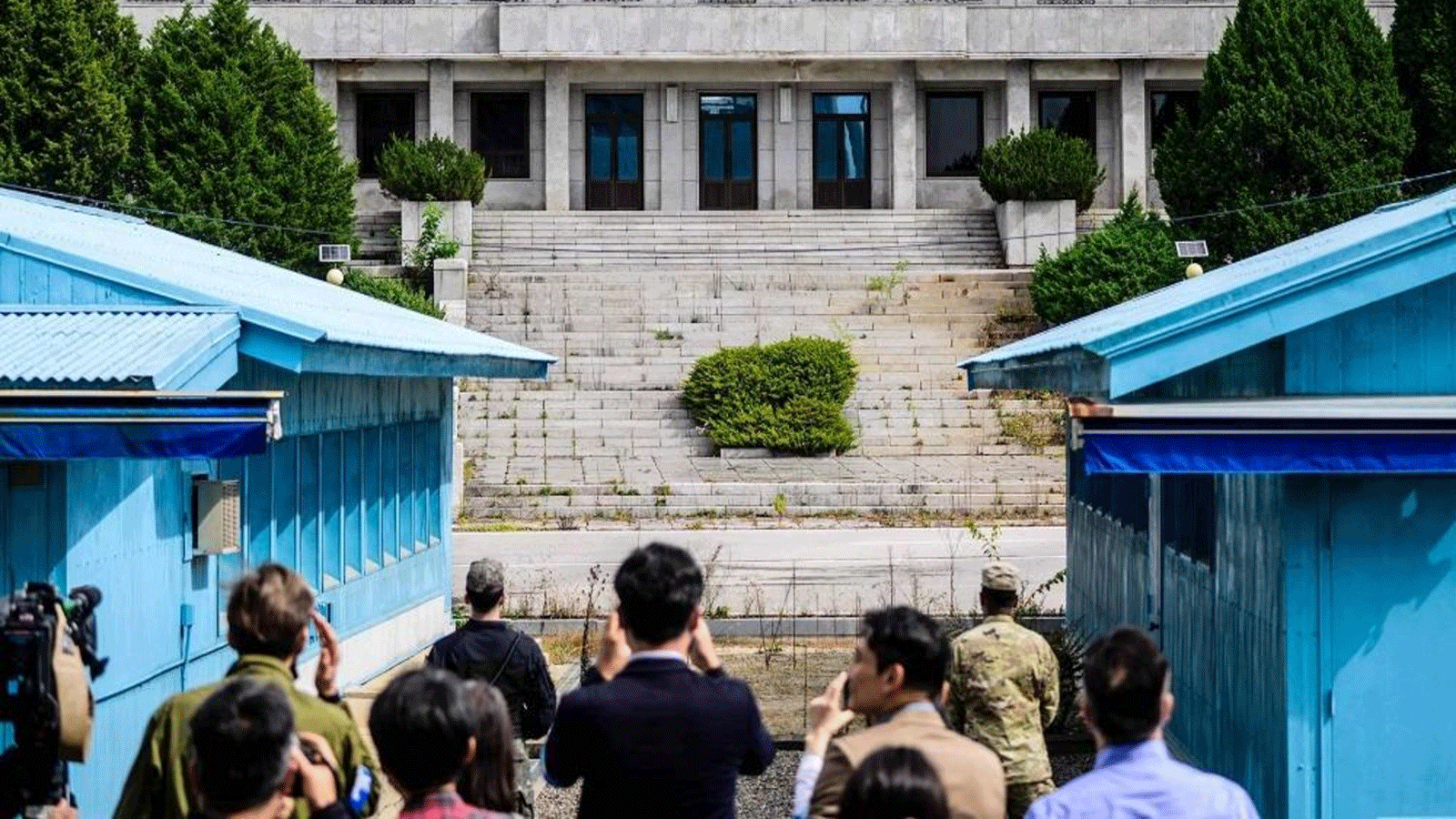 أعضاء وسائل الإعلام (في المقدمة) يلتقطون صورًا خلف جندي من قيادة الأمم المتحدة (مركز R) وجندي كوري جنوبي (الوسط L) أثناء وقوفهم في حراسة أمام قاعة بانمون في كوريا الشمالية (الجزء الخلفي C) وخط الترسيم العسكري الفاصل بين كوريا الشمالية وكوريا الجنوبية، في المنطقة الأمنية المشتركة (JSA) للمنطقة المنزوعة السلاح (DMZ) في قرية الهدنة، بانمونجوم.