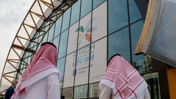 رجلان سعوديان أمام واجهة تحمل شعار إكسبو الرياض 2030