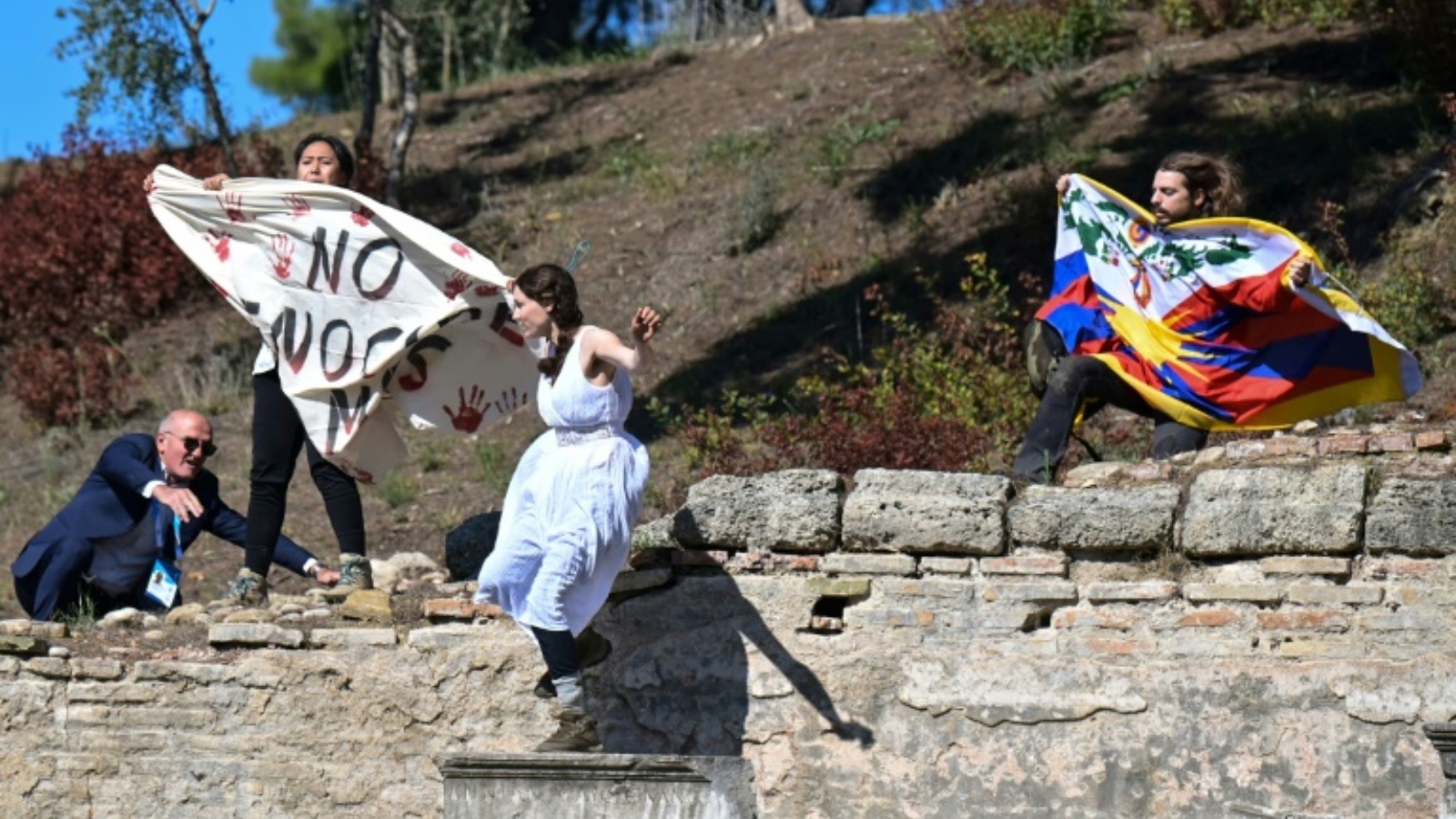 رجل أمن يحاول منع ناشطين من رفع علم التيبت ولافتة معارضة لاقامة اولمبياد بكين الشتوي لعام 2022، وذلك خلال إيقاد الشعلة الأولمبية في موقع أولمبيا القديم، جنوب اليونان في 18 تشرين الأول/أكتوبر 2021.