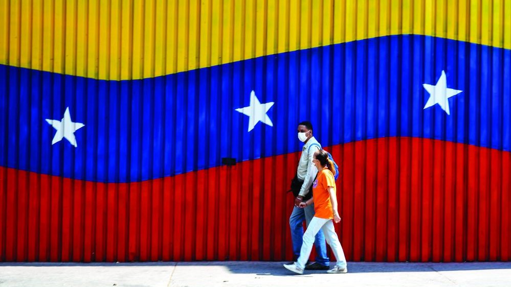 زوجان يرتديان أقنعة وجه يمشيان بجانب حائط رسم عليها العلم الفنزويلي في كاراكاس