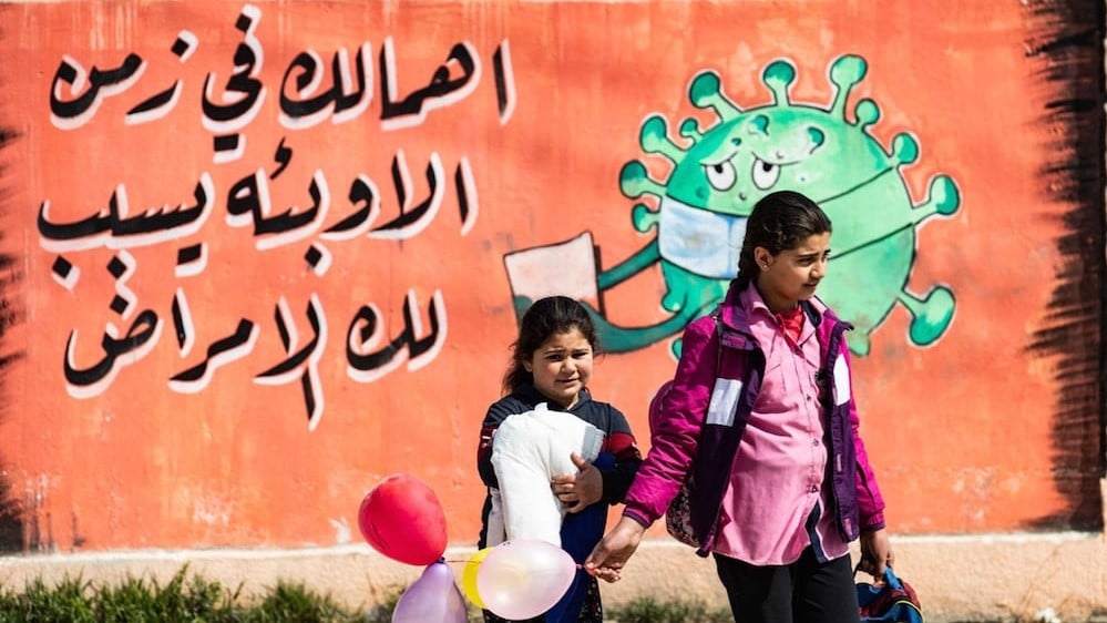 طفلتان تعبران أمام لوحة جدارية هي جزء من حملة توعية تدعو الناس إلى توخي الحذر أثناء الجائحة في القامشلي بسوريا، 9 مارس 2021