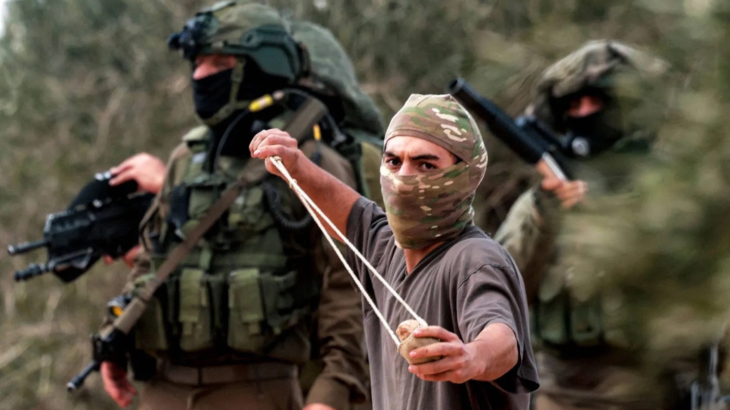 مستوطن ملثّم يلقي حجارة باتجاه فلسطينيين يقطفون الزيتون في الضفة الغربية بينما يقف خلفه جنود إسرائيليون