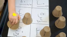 صورة تُظهر لوحة مكتوب عليها أحرفٌ باللغة العربية ويد طفل وهو يضع كوباً على كلمة إوزّة ضمن نشاطات حسّية لتعلّم اللغة العربية. - BBC