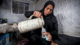 امرأة فلسطينية تصب الماء من إبريق كبير في زجاجة مخصصة لرضاعة الأطفال تحضيراً لإطعام طفل في خيمة. ترتدي المرأة حجاباً وعباءة سوداء. - Getty Images