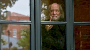 الكاتب الهنغاري لازلو كراسنهوركاي يقف خلف نافذة زجاجية، يرفع إصبعه بهدوء وينظر بتأمل إلى الخارج، فيما تنعكس خلفه ملامح مبانٍ من الطوب وأشجار خريفية. - Getty Images