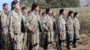 مجموعة من الرجال والنساء الأعضاء في حزب العمال الكردستاني، يحملون السلاح ويرتدون لباساً عسكرياً، يقفون في منطقة مفتوحة نهاراً. - AFP via Getty Images