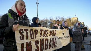 تبدو في الصورة فتاتان ترتديان ملابس شتوية، بشرتهما بيضاء، تحملان لافتة متوسطة الحجم مكتوب عليها باللغة الإنجليزية أوقفوا الحرب في السودان، وحولهما عدد من المتظاهرين. الصورة التقطت نهاراً والسماء زرقاء صافية. - Anadolu via Getty Images