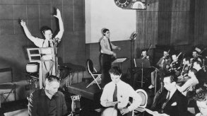 في استوديو إذاعة سي بي إس، يتدرب أورسون ويلز على أحد برامجه، واقفاً أمام الميكروفون مرتدياً قميصاً أبيض وربطة عنق داكنة، وذراعيه مرفوعتان بشكل درامي في منتصف العرض. على يمينه، يُجهّز أعضاء الأوركسترا آلاتهم الموسيقية، بينما يحمل الممثلون في المقدمة نصوصًا جاهزة لإشاراتهم. - Getty Images