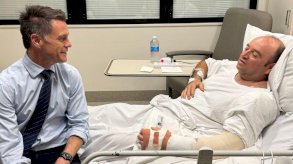 تُظهر الصورة مشهداً داخل غرفة في المستشفى. يرقد أحمد الأحمد على سرير مستشفى رمادي بملاءات بيضاء، وهو رجل أصلع يرتدي قميصاً أبيض، وقد وُضعت ذراعه اليسرى ومعصمه في جبيرة. ويجلس كريس مينز عند طرف السرير مبتسماً له، مرتدياً قميصاً أزرق فاتحاً، وربطة عنق زرقاء داكنة، وبنطالاً أسود. - Chris Minns/Facebook