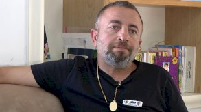 رجل يرتدي قميصا أسود وسروالاً كلاهما أسود اللون يجلس على أريكة لونها بني فاتح (بيج). شعره قصير داكن، وله شارب قصير ولحية قصيرة، تتخللهما خصلات من الشيب. يرتدي قلادة على شكل ميدالية ذهبية، وينظر إلى كرة قدم بيضاء على الطاولة التي أمامه، وقد طبع على الكرة وجوه وأسماء أطفال. - BBC