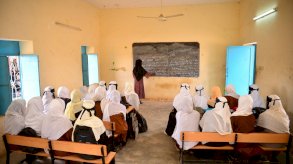 مجموعة من الفتيات تجلس داخل فصل دراسي بمدرسة في ولاية كسلا. وفي مقدمة الفصل، معلمة تكتب دروساً على سبورة وتشرحها. - Plan International / Abdelazim Yousif