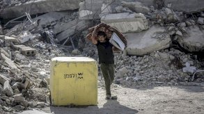 رجل يحمل مرتبة مطوية في غزة يمر بجوار كتلة خرسانية صفراء وضعها الجيش الإسرائيلي، وتظهر الأنقاض في الخلفية - Saeed M. M. T. Jaras/Anadolu via Getty Images