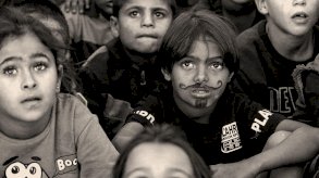 صورة بالأبيض والأسود لأطفال صغار يجلسون على الأرض وينظرون إلى الأعلى، وقد رُسم على وجه أحدهم شارب ولحية بالقلم. - BBC