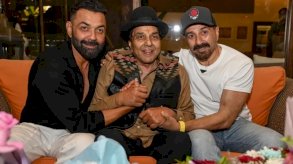 النجم دارمندرا (في الوسط) مع ولديه ساني (يمين) وبوبي ديول (يسار) في مقر إقامته في مومباي. دارمندرا بعد أن كبر في السن ويرتدي قبعة سوداء وقميصاً عسلي اللون وجاكيت بدون أكمام أسود وبنطالاً أسود في المنتصف بين ابنيه الشابين أحدهما (ساني) يرتدي قميصاً أسود والآخر قميصاً قصير الأكمام بلون أبيض. - AFP via Getty Images