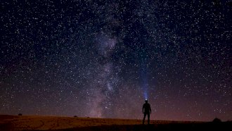 ظلّ إنسان يقف تحت سماءٍ مترامية، يرفع نظره نحو مجرّة درب التبانة وهي تشقّ الظلام. - Anadolu via Getty Images