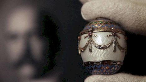 موظف في دار كريستيز يحمل علبة هدايا بيضة ذهبية ثنائية اللون من مينا فابرجيه أمام صورة للملك اليوناني جورج الأول. البيضة مزينة بتفاصيل ذهبية، مركزها أبيض، وتفاصيل بنفسجية وخضراء في الأعلى والأسفل. - Getty Images